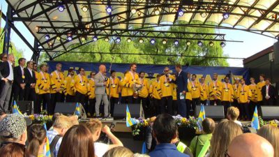 Det svenska VM-laget står i sina hockeytröjor uppe på en stor scen i Kungsträdgården i Stockholm. I bakgrunden syns träd och framför scenen står människor med svenska flaggor.