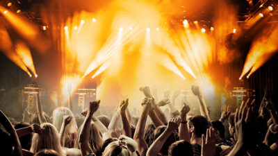 Människor som står samlade framför en stor scen. Strålkastarna lyser och publiken håller upp sina händer.